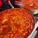 Large pans of spicy Korean tteokbokki cooking at a street food stall with red sauce and rice cakes