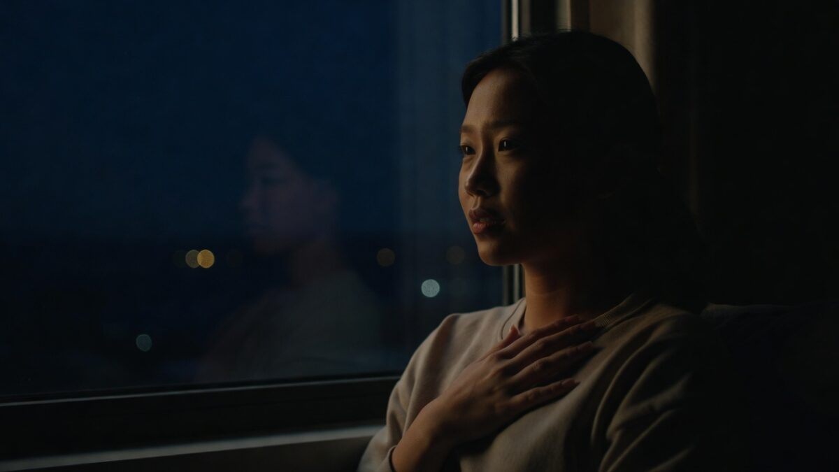 Person sitting alone by a window at night pressing hand to chest with soft hopeful expression